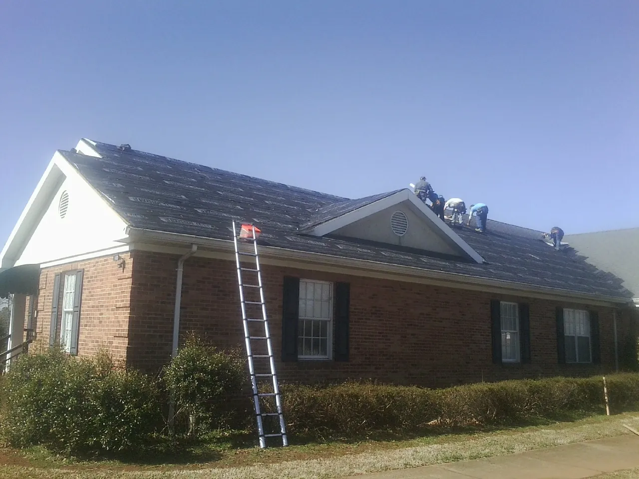 Crew tearing shingles off a brick ranch roof with a ladder set — Metal Roof Repair work by our crew in Lake Ariel